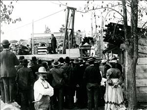 A crowd of people stand around an outdoor stage. On the stage a man stands in front of a box draped in an American flag. 